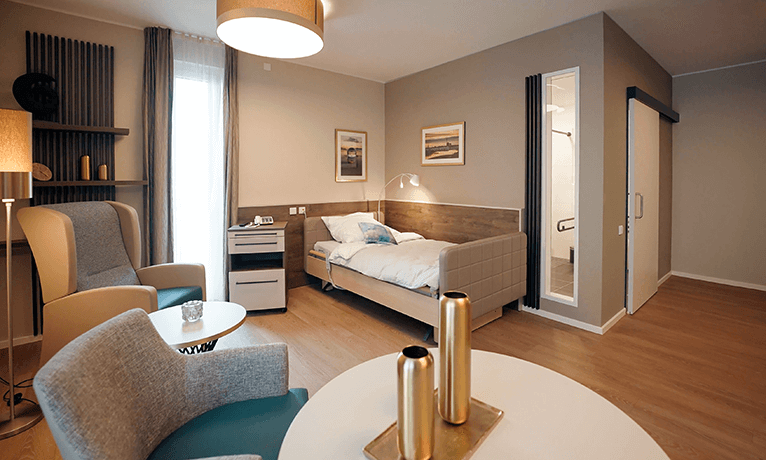 Individuelles Einrichtungskonzept mit einem Bett, einem Nachtschrank, einem Wohnzimmertisch in rund und einem kleinen Sofa in einem großräumigen Bewohnerzimmer.
