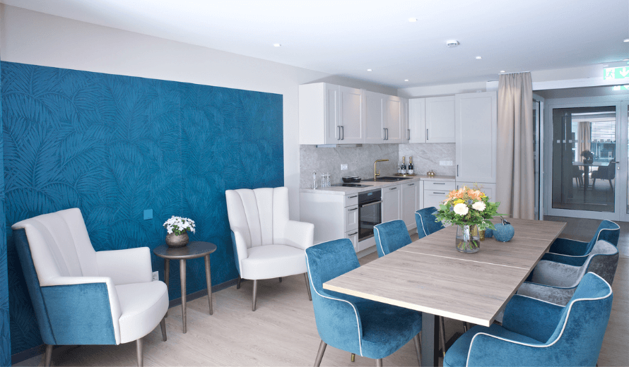 Eingerichtetes Bewohnerzimmer in blau weiss und einer Küche, Esstisch und Stüheln.